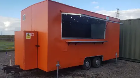 A large orange trailer is parked on some gravel. A serving hatch is open showing ceiling lights inside the trailer. It has wheels in the centre of the trailer and a tow bar at the end, on the left of the picture