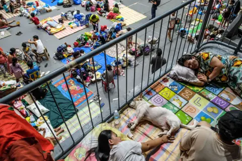 EPA Lots of families sleep on the floor in what looks like a large hall, with other families gathered around with rugs and posessions on the floor.