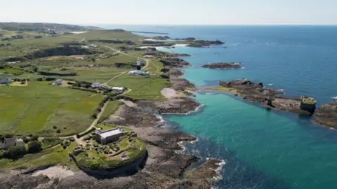 An aerial view of some of the Alderney coast. It shows green fields, some rugged coastline, a lighthouse, and what appears to be some fortifications on the island and on an islet.