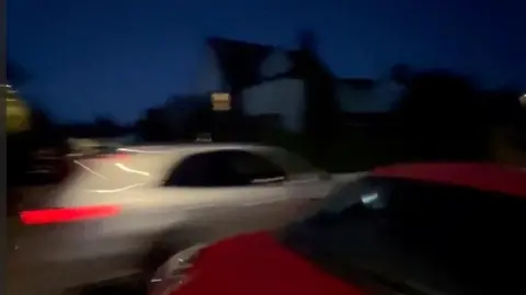 Supplied A blurred night-time scene capturing a fast moving white car on a road, speeding past a stationary red car. Street signs and silhouettes of trees appear against a dark blue evening sky.