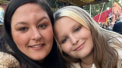 Family handout/PA A young girl with blonde hair rests her head on the shoulder of her mum, who has dark hair. Both are smiling into the camera.