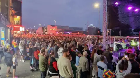 A large group of people stand in front of the main stage at dusk in the town.