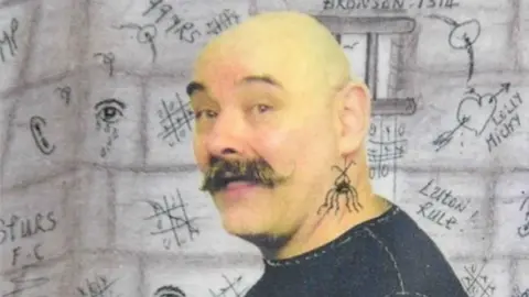 Charles Bronson, now known as Charles Salvador, is bald and has a moustache. He's wearing a black top.