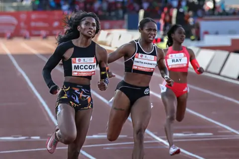 TONY KARUMBA / AFP / GETTY IMAGES Three women run across the finish line on an athletics track. Two on the left are dressed in black and represent Liberia. The woman to their right is dressed in red and represents the USA.