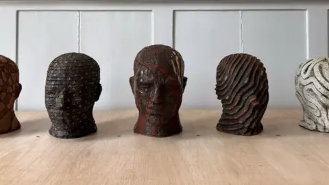 Sketches and maquettes of Joseph Hillier's head busts for the art train in Watlington. All five of them have different designs and are placed on a wooden table in a room.
