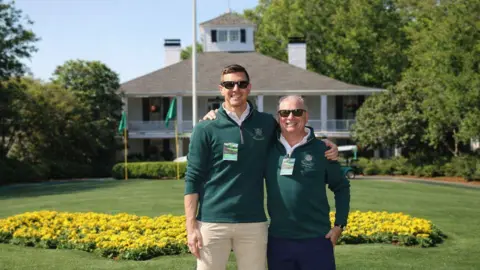Gareth Maher Two men standing outside the club house at the golf course. Both men are wearing green jumpers and sunglasses. The man on the left has brown hair and the man on the right has grey hair. In the background, you can see the white club house with grey roof and lots of trees with green leaves. There's also a flower bed of yellow flowers behind them.