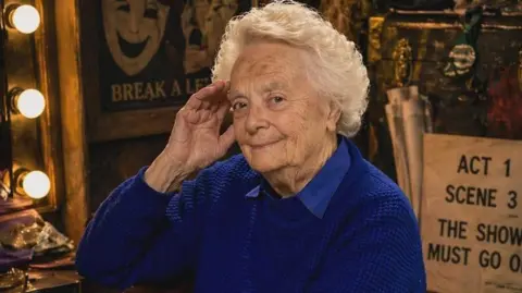 A 92-year-old woman with short curly white hair sits at a dressing table surrounded by theatre props. She is dressed in a blue pullover.