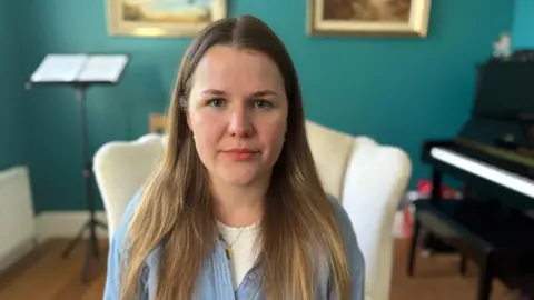 Supplied A woman with long, brown-blonde hair looks directly into the camera with a neutral expression. She is wearing a blue top with a white top underneath. In the background a piano and a music stand can be seen, as well as a turquoise-coloured wall and cream armchair.