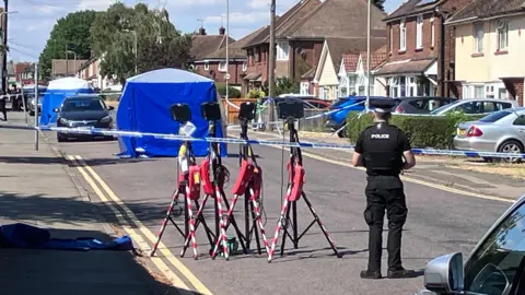 A blue police tent is set up in the middle of a residential street. Police tape blocks the road. Another tent is set up further along the road. A police officer stands with his back towards the camera as he faces the scene.