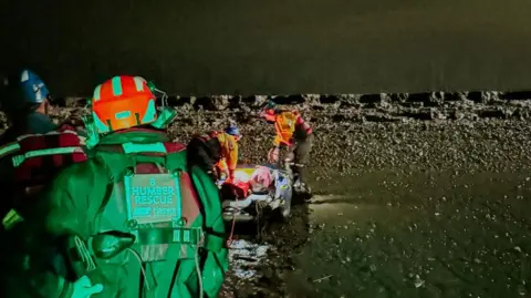 The Humber rescue team attend a casualty, who cannot be seen, on the the edge of the estuary.  The team wear helmets and hi-vis suits with "Humber Rescue" written on them. The scene is illuminated at night, revealing a muddy shore and dark water. 