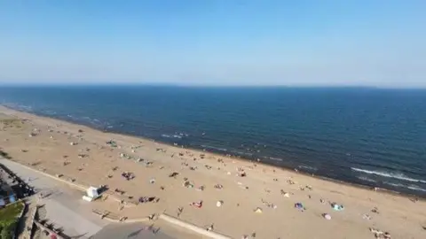 Sandy beach with people sitting on it. There is a blue sea and blue sky.