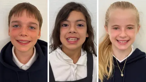 A composite of three school children. Sam is a boy with short brown hair, he is smiling and wearing a dark hooded top and white shirt. Grace has long dark brown hair and is smiling widely. She's wearing a white shirt and grey dress. Betsy has long blonde hair in a ponytail over one shoulder. She is smiling and waring a daisy necklace  over a dark hooded top