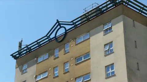 Top of a tall block of flats underneath a blue sky. Decorative iron work on top and rendered in a cream and sand colour.