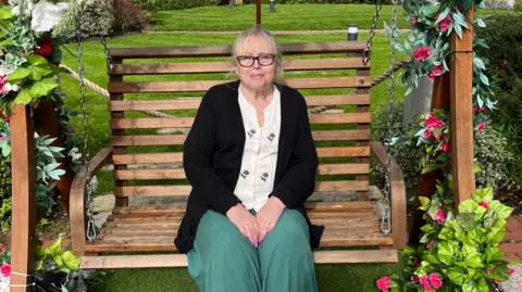 Hazel Busby Hazel Busby sitting on a wooden swing bench surrounded by flowers