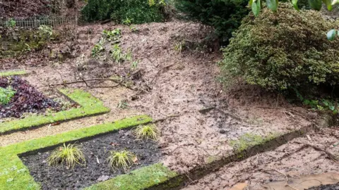 Thick mud covers parts of a previously neat, landscaped garden. Some plants can be seen emerging from the soil.