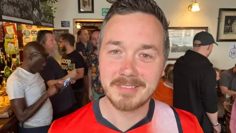 Sam Read/BBC Robin Wheeler in the pub with punters at the bar behind him. It is busy and people are drinking and chatting. He wears a football shirt.