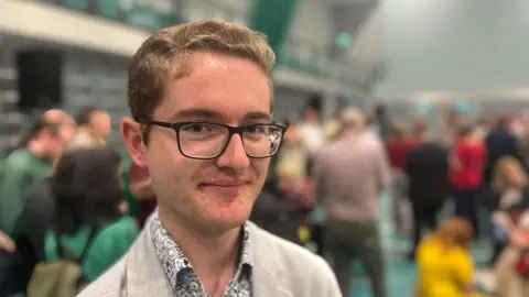 A picture of Thomas Richardson, 20-year-old Green party candidate elected to Devon County Council. He is smiling while wearing a grey suits and having black glasses.