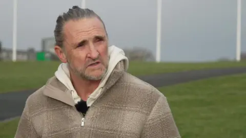 Paul Maxwell, wearing a beige fleece and hoodie, looks towards the right of frame. He is sat on a bench on Plymouth Hoe.