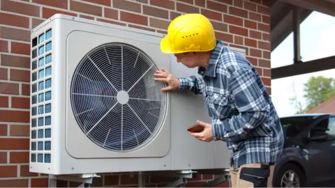 A man in a yellow hard hat examining an external heat pump on the wall of a house. The pump is a large grey box with a fan in it.