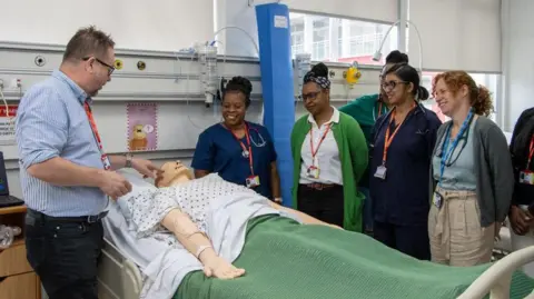 University of Bedfordshire A man and a group of women stand over a mannequin lying in a hospital bed. There are five women standing over the mannequin.