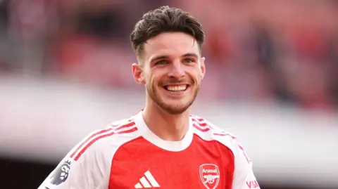 PA Media Declan Rice in an Arsenal football shirt. He is smiling.