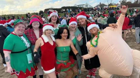 Eight woman stand and smile at a camera. They are all wearing red Santa hats, that have white headbands. The woman on the far right wears a blow up peach-coloured Turkey costume.