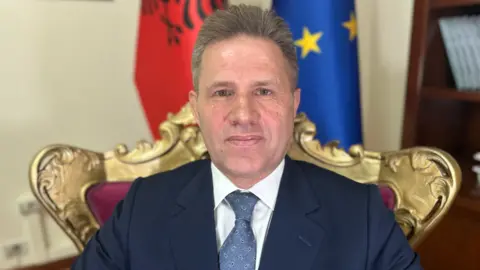 Josie Hannett / BBC Lavdrim Krashi sitting inside the government building in Tirana, the Albanian capital, on a chair in front of flags for Albania and the European Union