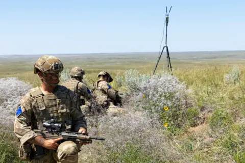 Crown Copyright 2026 A soldier in green uniform and protective gear faces the camera, standing or kneeling among green bushes. Behind him are two men in green and brown camouflaged uniform facing the other way. A large expanse of green fields can be seen in the background. A black tripod has been placed nearby.