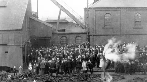 Beamish Museum Historic black and white photograph showing the aftermath of the West Stanley Colliery disaster in 1909. A crowd, mostly composed of men are milling around in front of tall, brick colliery buildings. Debris - what appears to be metal - is to the foreground and to the right a pipe is emitting smoke or steam.