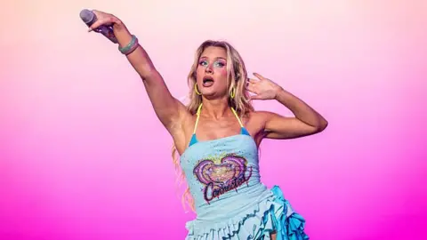 BBC Zara Larsson performs on stage and she is holding a microphone outstretch to the crowd. She wears a blue sparkly dress.