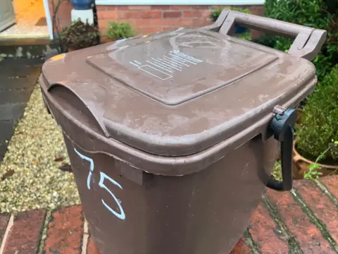 A brown food waste bin with the number 75 painted on the side sits on a red brick wall in front of a house. 