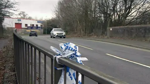 BBC Police tape tied on to a metal bridge on Dartmouth Road, an industrial street lines with trees and bushes