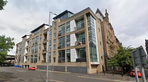 A modern multi‑storey apartment building with large glass windows and stone cladding on a quiet city street corner. A few parked cars and road signs sit in the foreground, with older historic buildings visible to the right and cloudy sky overhead.