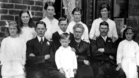 Tarran family A family group picture of 11, taken outside a brick house with a window with lace curtains in the background. The picture was taken around 1918, so all the people are wearing Edwardian style formal clothing, suits for the men and lace dresses for the girls. There is also a woman wearing a dark silk dress. Three of the family, a woman and man who appear to be middle aged, and a younger man are seated, and the rest of the family, all children and young people are standing to their side or behind them. One young woman has her hands resting on the older man's shoulders. The youngest child, a boy of 6, is standing next to the older woman wearing a white sailor suit style outfit. The boy behind him has his hand on his shoulder.