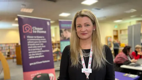 Laura looks at the camera as she stands in the Library with banners up behind her and people sitting down and talking. She is wearing a black jumper and has long blonde hair which comes over her shoulder.