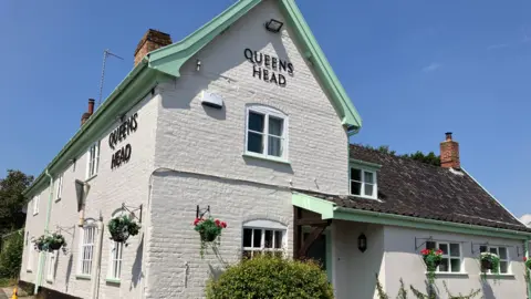 GUY CAMPBELL/BBC Painted white brick facade of the Queens Head pub with flowers in baskets hanging from the walls