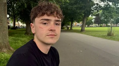 Charlie Weatherley sits next to a path in a public park, facing the camera. He wears a black T-shirt with white printed text. Behind him, a wide grassy area stretches out with trees, houses and parked cars visible in the distance. The path curves slightly and is bordered by grass on both sides.