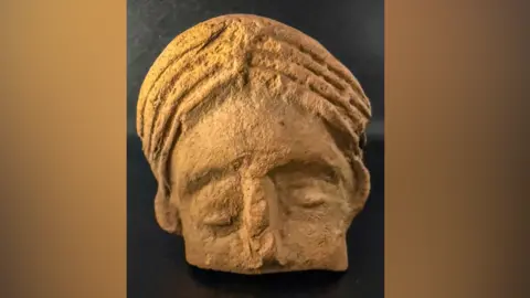 The terracotta head of a woman has been found. The eyes are not symmetrical and the hair shows signs of wear. The bottom half of the fact below the nose is missing.