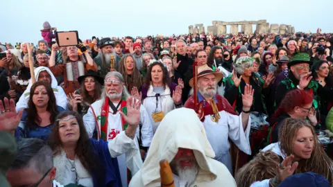 PA Media A large crowd of people dressed in ceremonial clothes like cloaks, headbands, face paint and ponchos. They are all looking towards the sunrise with their hands held in the air. In the background you can see Stonehenge against the pale blue sky.