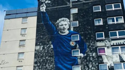 High-rise building with mural of late footballer Denis Law with his arm aloft, in a blue Scotland shirt.