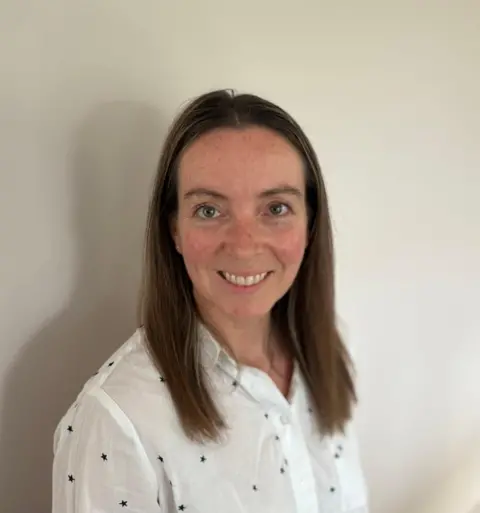 East West Rail A picture of a woman who is stood in front of a plain white background. She has brown shoulder length hair and is wearing a white shirt with black dots on it.