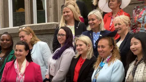 A large group of woman stood together. They are wearing various colourful smart outfits.