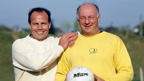 Coventry City Football Club George Curtis and John Sillett: Curtis stands with his hands on Sillett's shoulders. Both smile at the camera as Sillett holds up a football