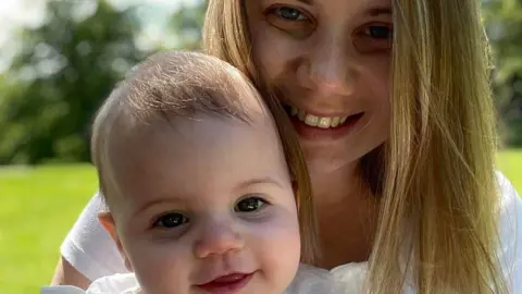 Family handout Rebecca Ableman is smiling while holding her daughter, Autumn. They are both outside on the grass during a sunny day and are wearing white. Autumn is smiling and looking directly at the camera. Rebecca is doing the same and has long blonde hair. 

