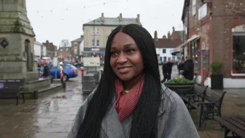 Nnenna Hemeson, who has long black hair and is wearing a grey coat with a pink scarf.