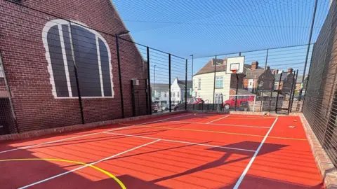 Supplied A red outdoor sports court with white and yellow lines and a basketball ring at one end.