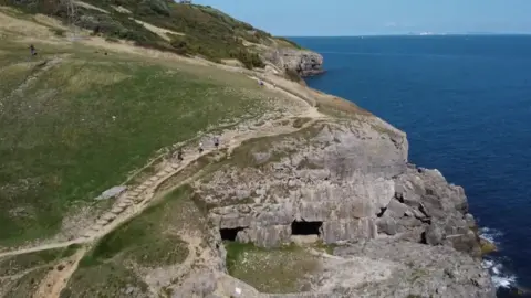 A drone shot of a path snaking its way across the edge of a coastal cliff. A few walkers can be seen ascending some stairs on the route. Down at the foot of the cliff, ocean waves can be seen splashing against the rocks. 