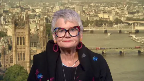 Carolyn Harris MP is wearing brightly-patterned glasses, large purple hoop earrings, silver necklaces and a black top and jacket with blue and purple embroidery. She is sitting in front of a backdrop of the Houses of Parliament and the River Thames.