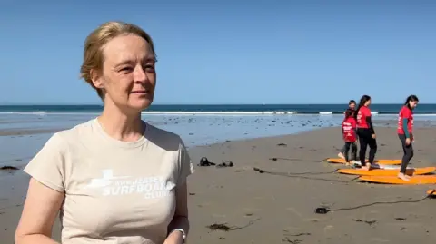 Michelle is wearing a light-coloured T-shirt stands on a sandy beach in the foreground, facing the camera. In the background, four people in red shirts practice balancing on orange surfboards laid out on the sand during a surf lesson. The beach stretches to the shoreline under a clear blue sky, with a historic stone tower visible on a small rocky islet in the distance. 