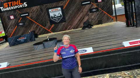 A woman with blonde hair, wearing a red and blue T-shirt, black trousers, a watch and a medal, stands in front of a wooden stage.